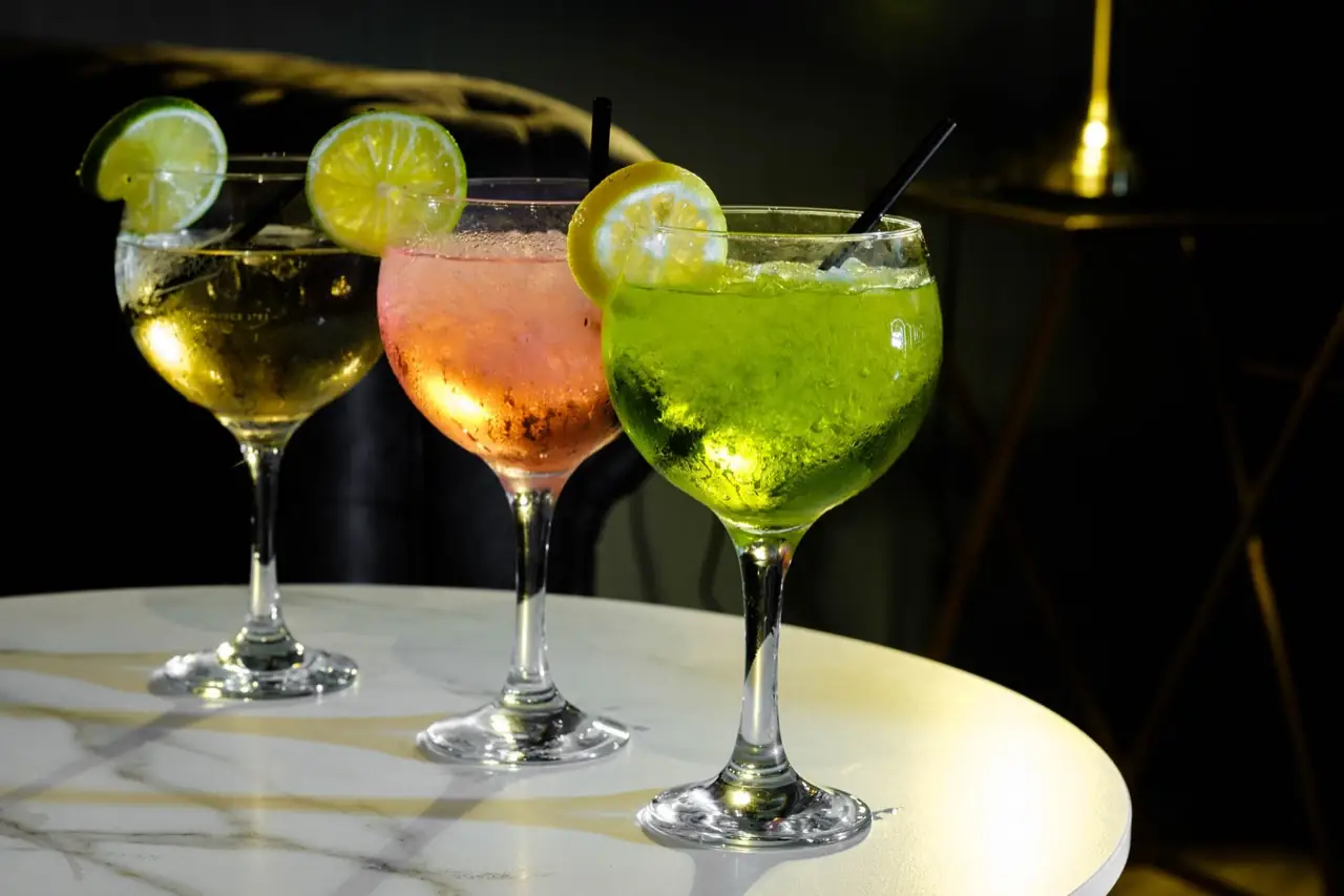Three balloon-glass cocktails (yellow, pink and green) garnished with lemon and lime slices on a white marble table in a dimly lit bar