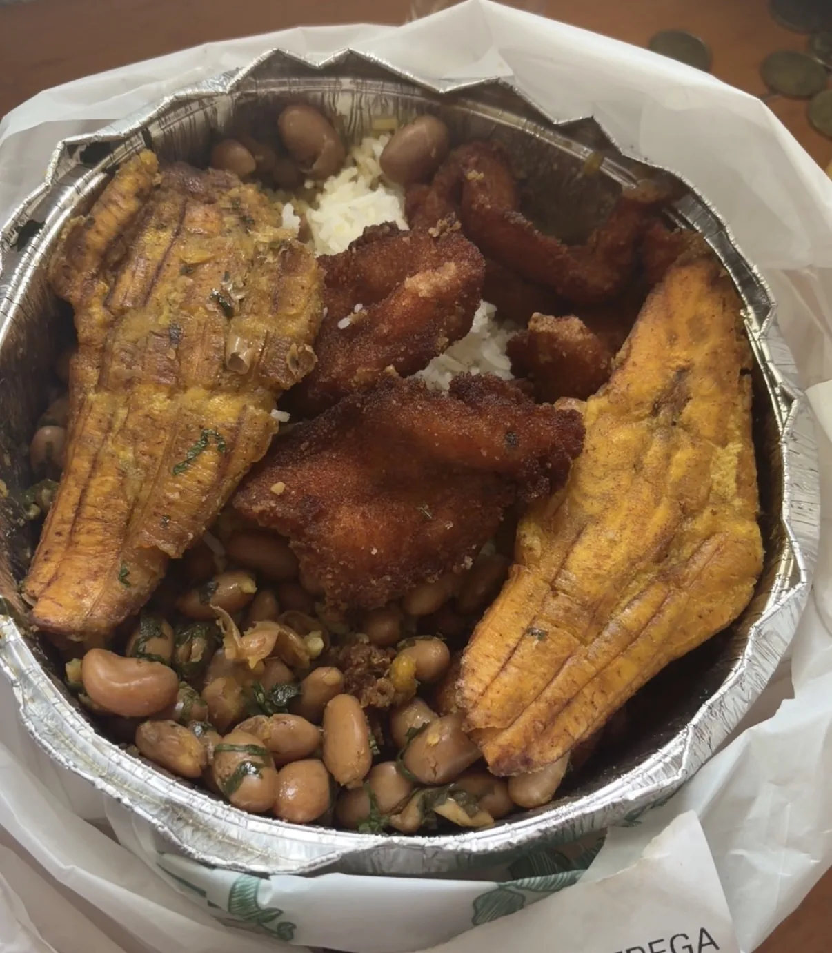 Fried plantains and pork served in a foil container alongside a side of beans