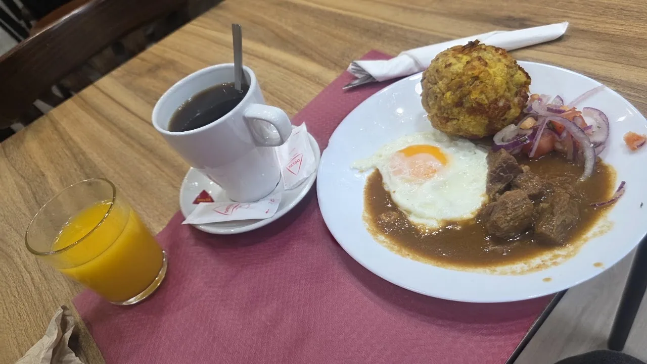A breakfast plate with a fried egg and a croquette in sauce, next to a cup of coffee and a glass of juice