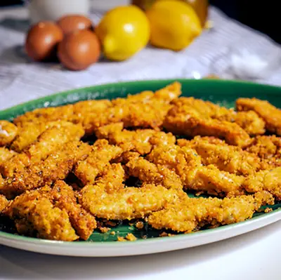 Green oval platter piled with breaded fried chicken strips, with brown eggs and lemons in the background
