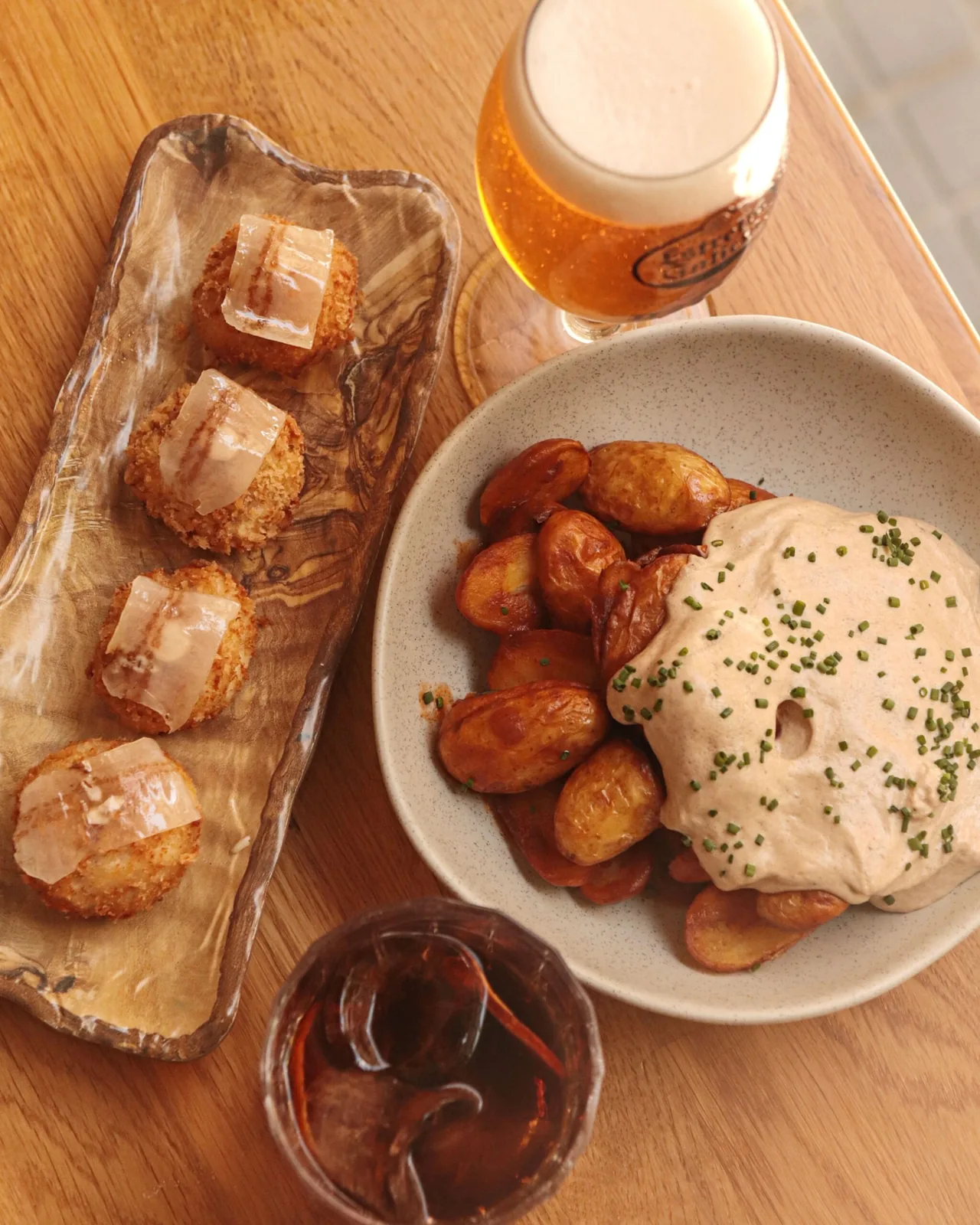 Four breadcrumbed croquetas topped with a thin veil of cured fat on a wooden dish beside a glass of beer and a bowl of potatoes