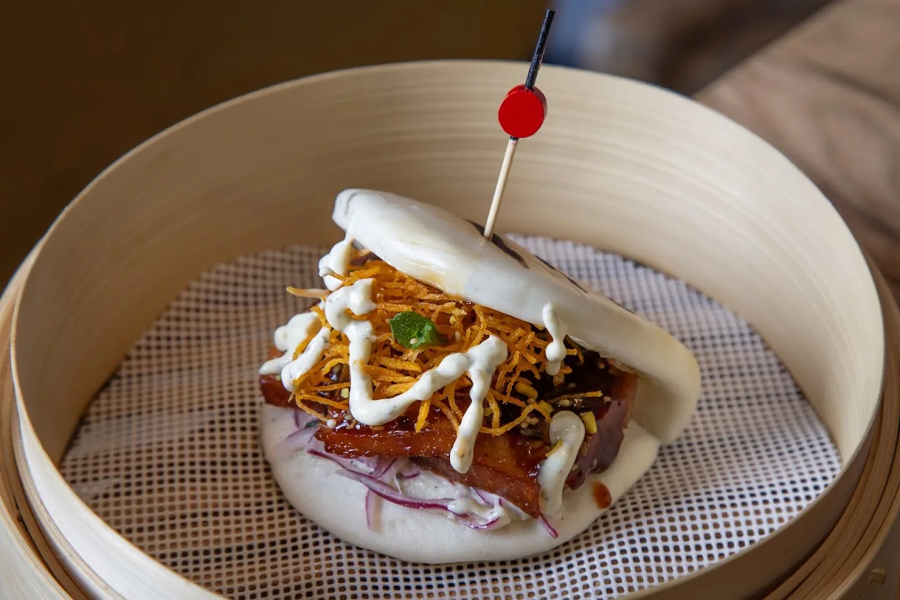 A steamed bao bun filled with meat and drizzled with sauce, on a skewer