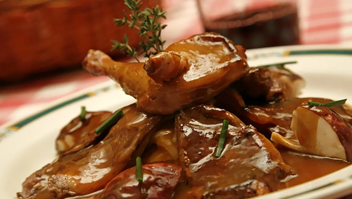 A braised poultry leg on sliced potatoes in glossy brown sauce, garnished with a thyme sprig and chives