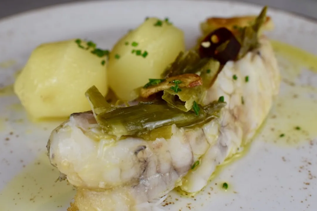A white fish fillet with green sauce, boiled potatoes, and garlicky leek strips on a white plate