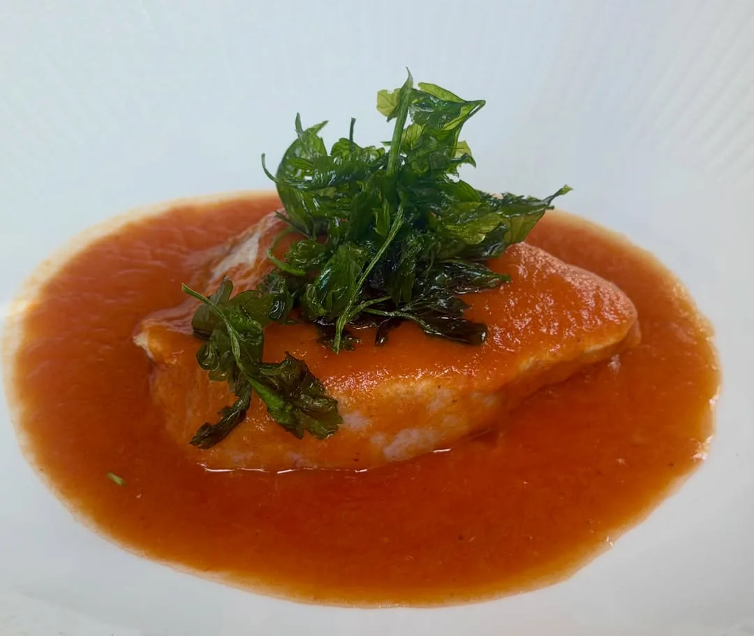 A pan-seared fish fillet in tomato sauce, topped with fried herb leaves, on a white plate