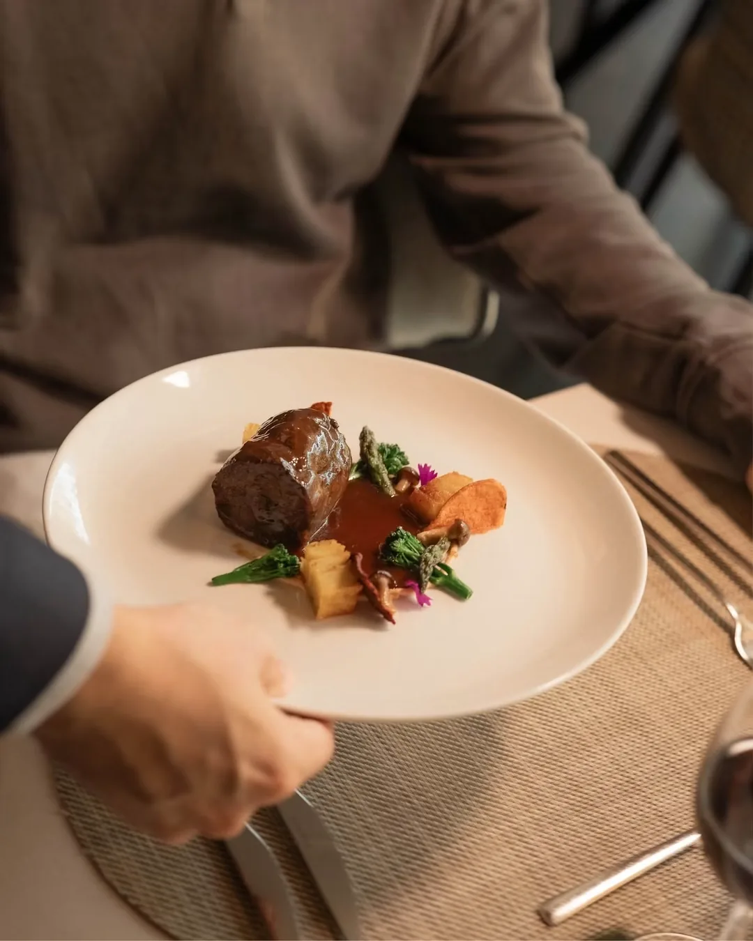 A small plated dish with a piece of roasted meat, orange puree, and microgreens, held by a server