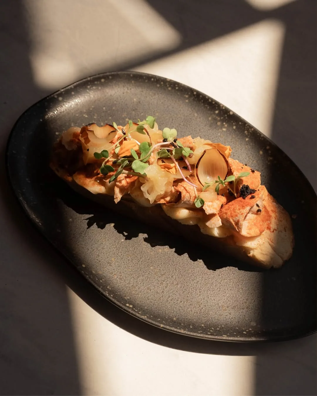 An oval dark plate with a crostini-style toast topped with shrimp, thin apple slices, and herbs