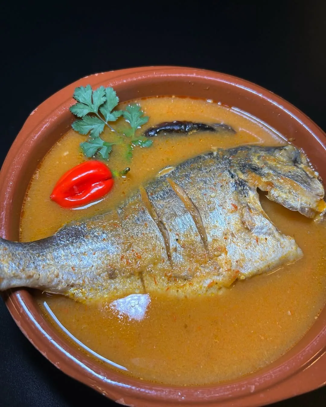 A whole fish in an orange broth in a clay bowl with a red pepper and herbs