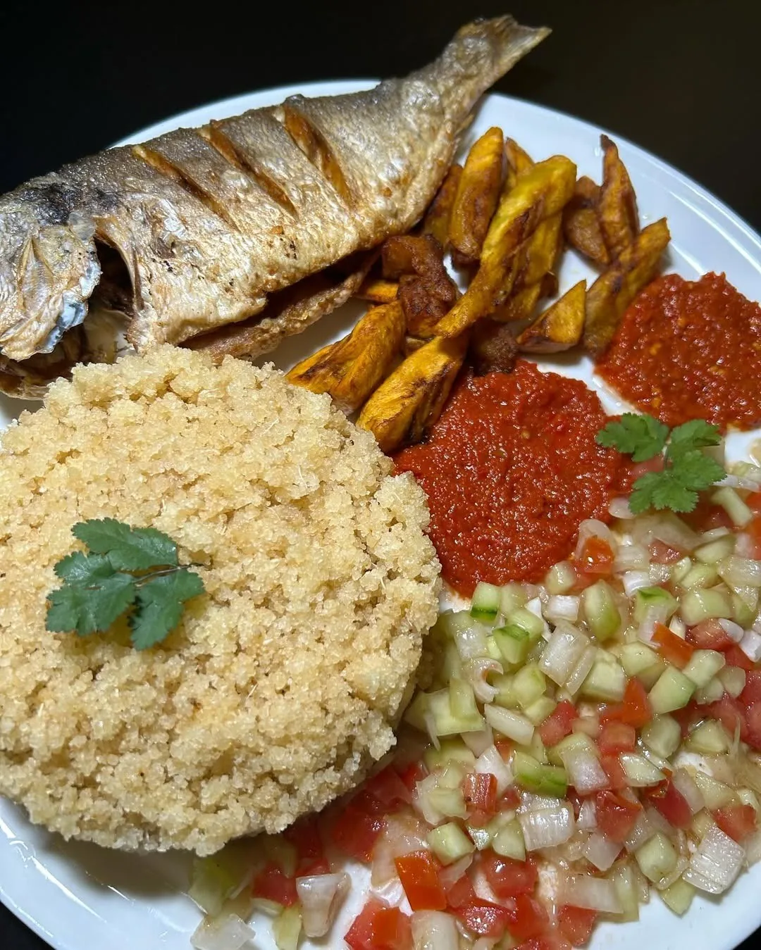 A plate with a grilled fish, fried plantain, couscous, red sauce and chopped salad