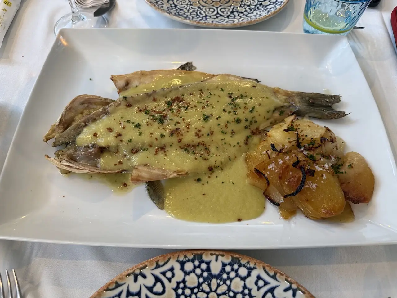Whole grilled fish in a green herb sauce with roasted potatoes on a square plate.