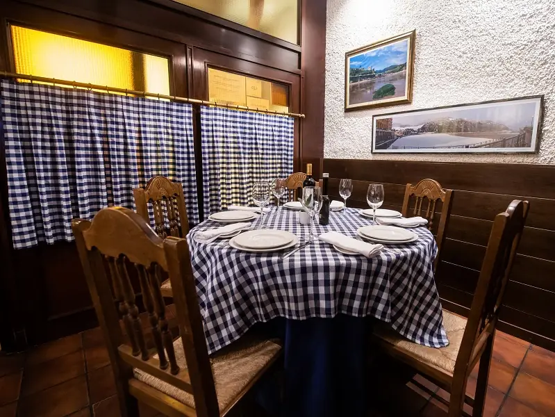 Traditional dining nook with a round table set with blue-and-white gingham cloth, white plates and wine glasses, wooden chairs and framed river photographs on a textured wall
