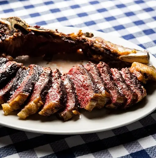 Sliced grilled T-bone steak showing a rare pink center on a white platter set on a blue-and-white gingham tablecloth
