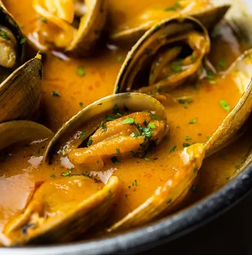 Close-up of clams in a saffron-orange sauce garnished with chopped parsley in a dark bowl