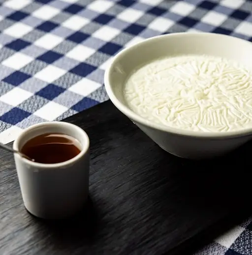 White bowl of cuajada or fresh curd next to a small cup of dark syrup on a black board over a blue gingham cloth
