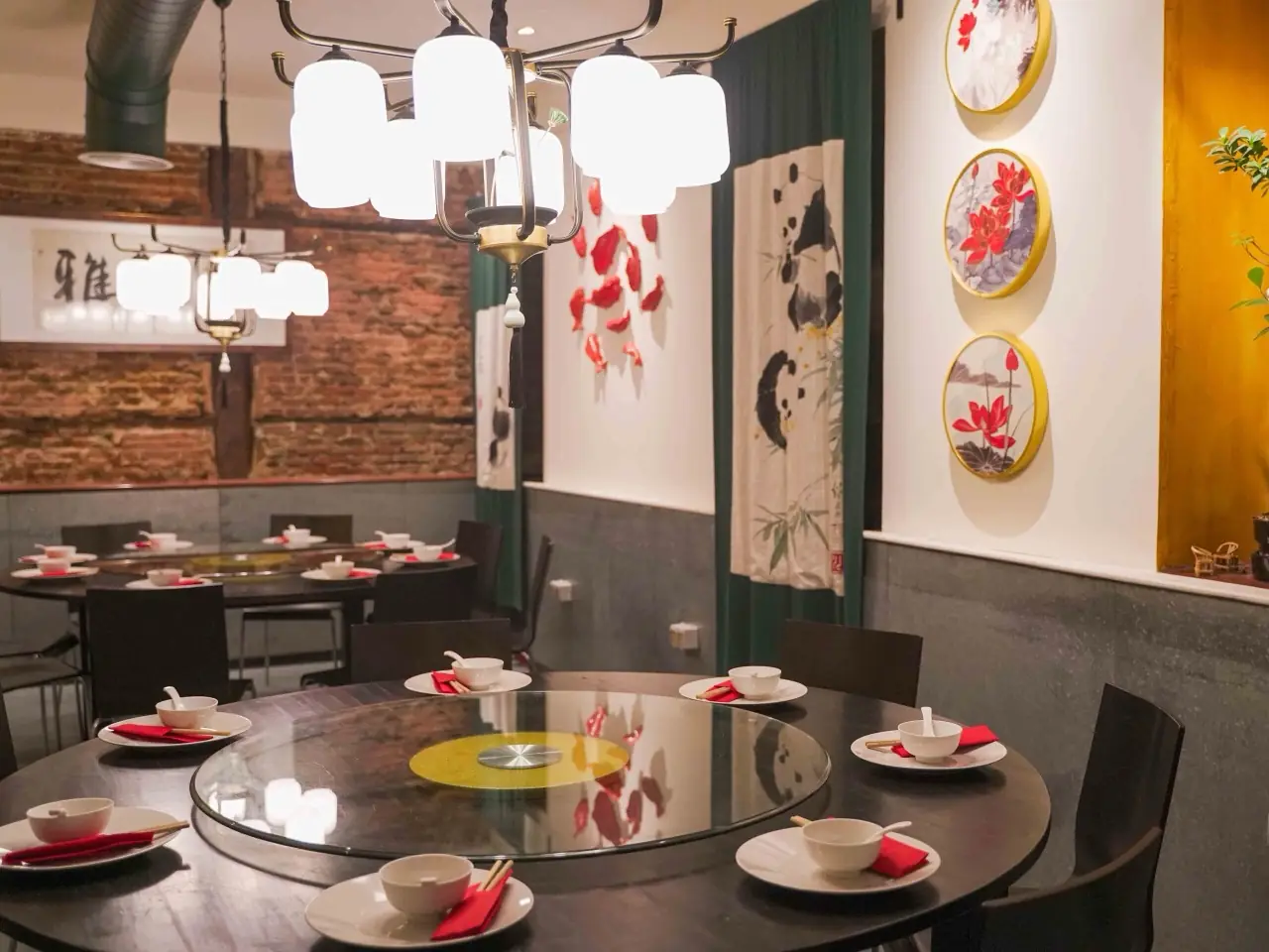 Chinese restaurant dining room with round tables under glass lazy Susans set with white bowls and red napkins, exposed brick, globe chandeliers and panda-and-lotus wall art