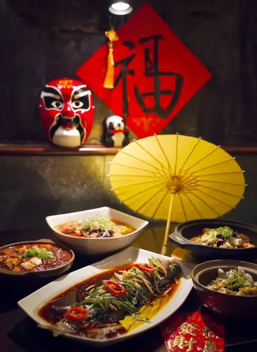 A spread of Sichuan dishes including a whole chili-braised fish and clay-pot dishes, with a red Peking-opera mask, panda figurine and yellow paper parasol as decor