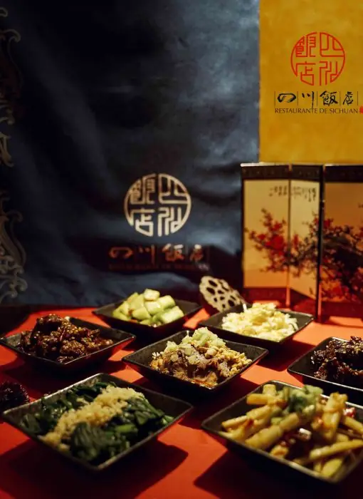 An array of small square dishes of Sichuan appetizers on a red cloth in front of a blue banner bearing the Restaurante de Sichuan name and seal