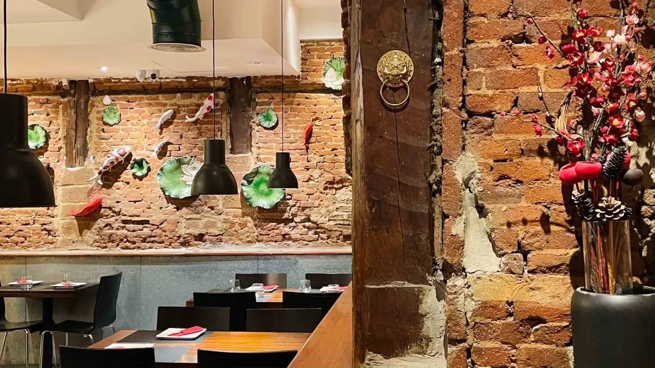 Restaurant interior with an exposed brick wall decorated with ceramic koi and lotus leaves, black pendant lamps, a carved timber column with a brass lion door-knocker and a vase of red blossom branches