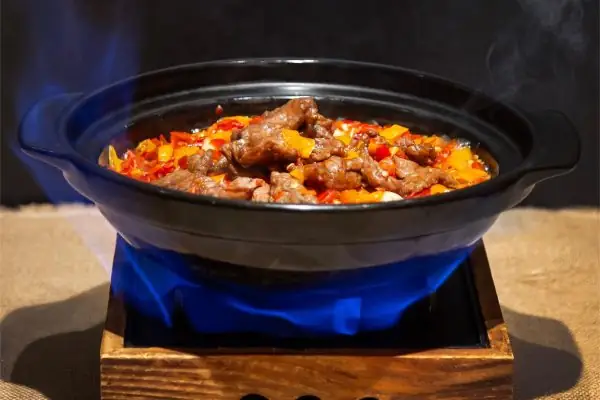 A black clay pot of sizzling stir-fried beef with red and yellow peppers over a blue flame on a wooden stand