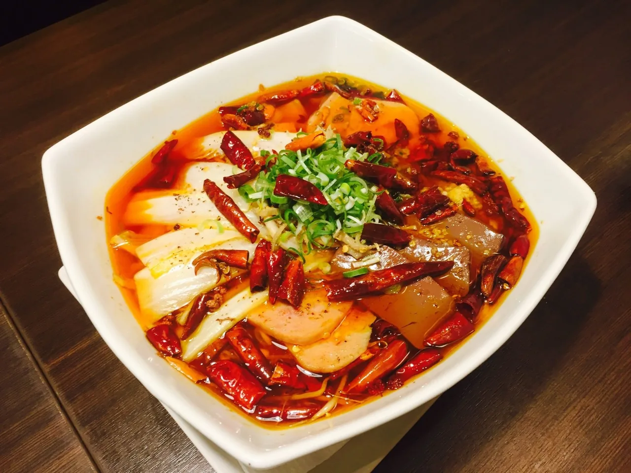 A white bowl of red chilli broth with dried chillies, spring onion and sliced ingredients