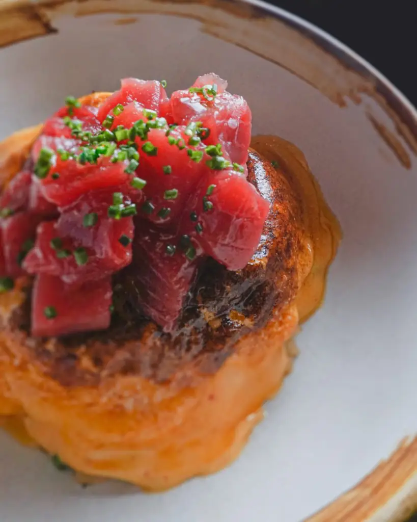 Diced raw tuna tartare sprinkled with chopped chives, mounded over a seared savory cake in a rustic bowl