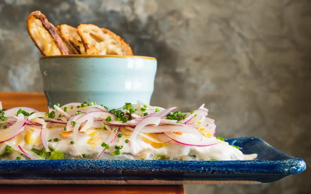 Cured fish dish topped with sliced red onion and chives on a blue plate, with grilled bread in a pale bowl