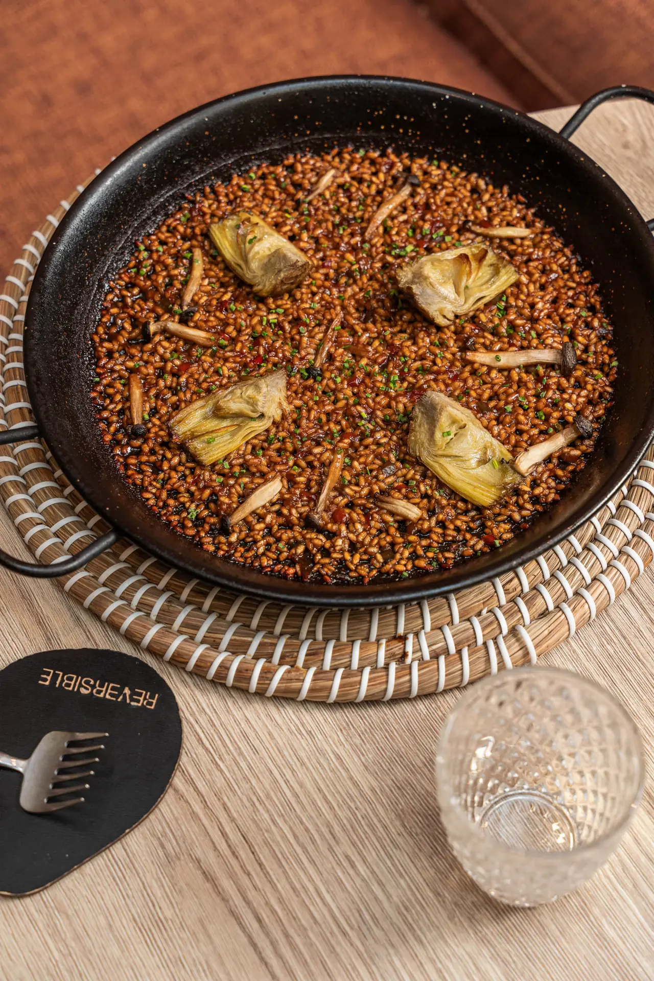 Rice dish with grilled baby artichokes and shaved mushrooms in a black paella pan, garnished with chives, Reversible Barcelona