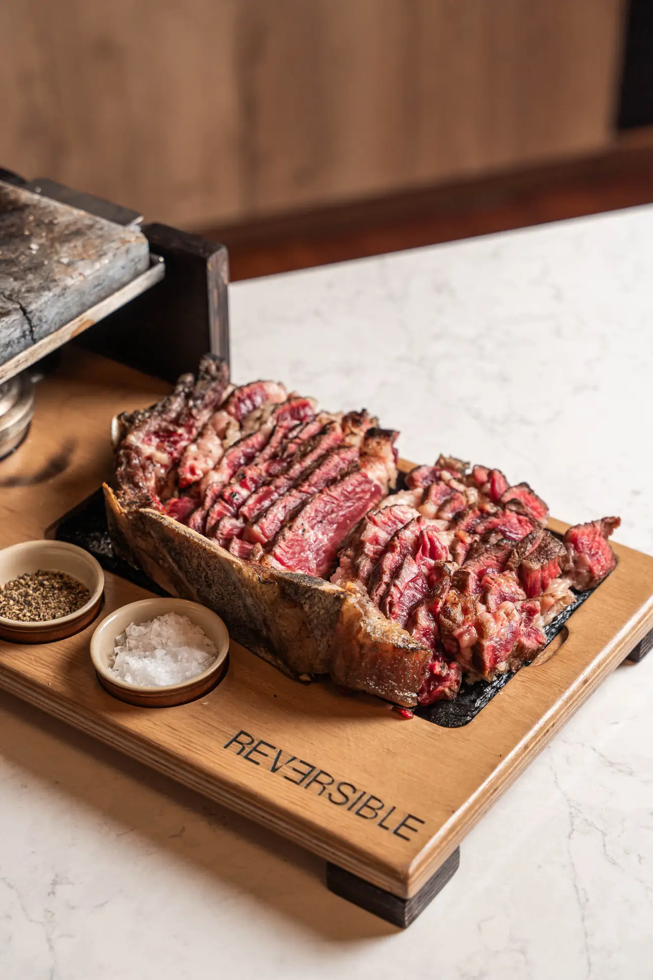 Thick-cut steak sliced into strips on a wooden board with bone attached, served with small bowls of salt and pepper, Reversible Barcelona