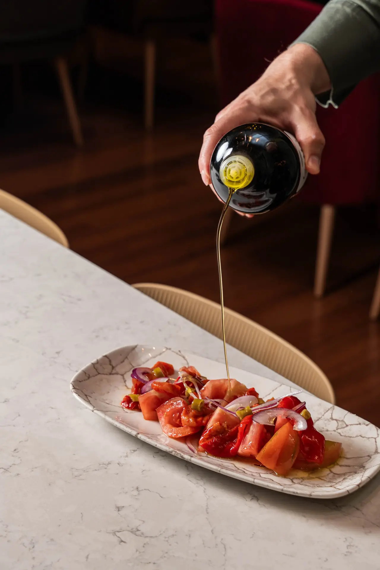 Chopped tomato salad with sliced red onion and green pickles on an oval plate, being drizzled with olive oil, Reversible Barcelona