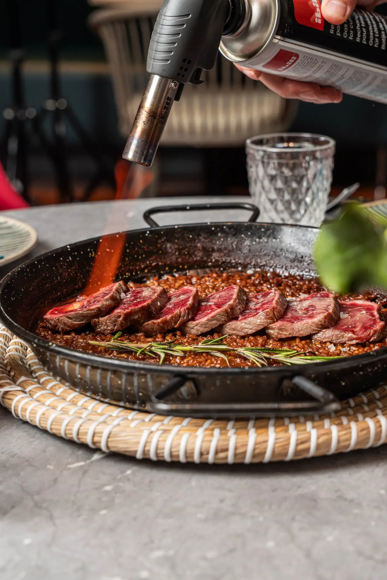Sliced steak medallions over rice in a metal pan, being finished with a kitchen torch and rosemary sprigs, Reversible Barcelona