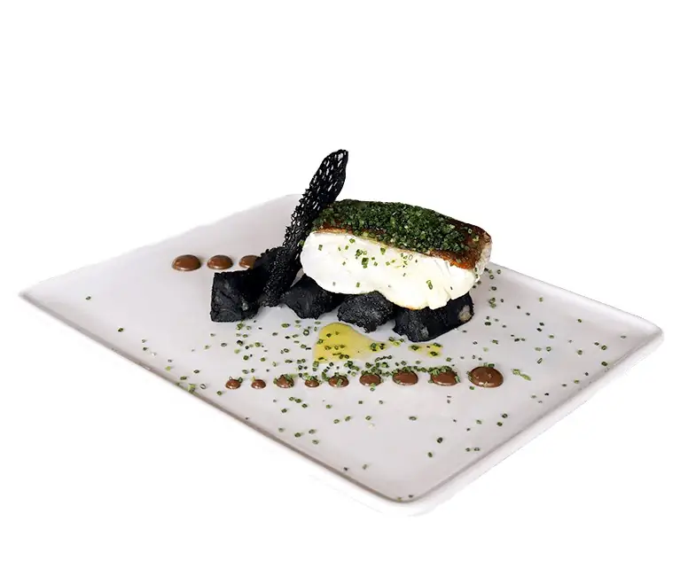 Plated white fish fillet with a green herb crust set on black-hued garnish, with dabs of brown sauce and chive scatter on a white rectangular plate
