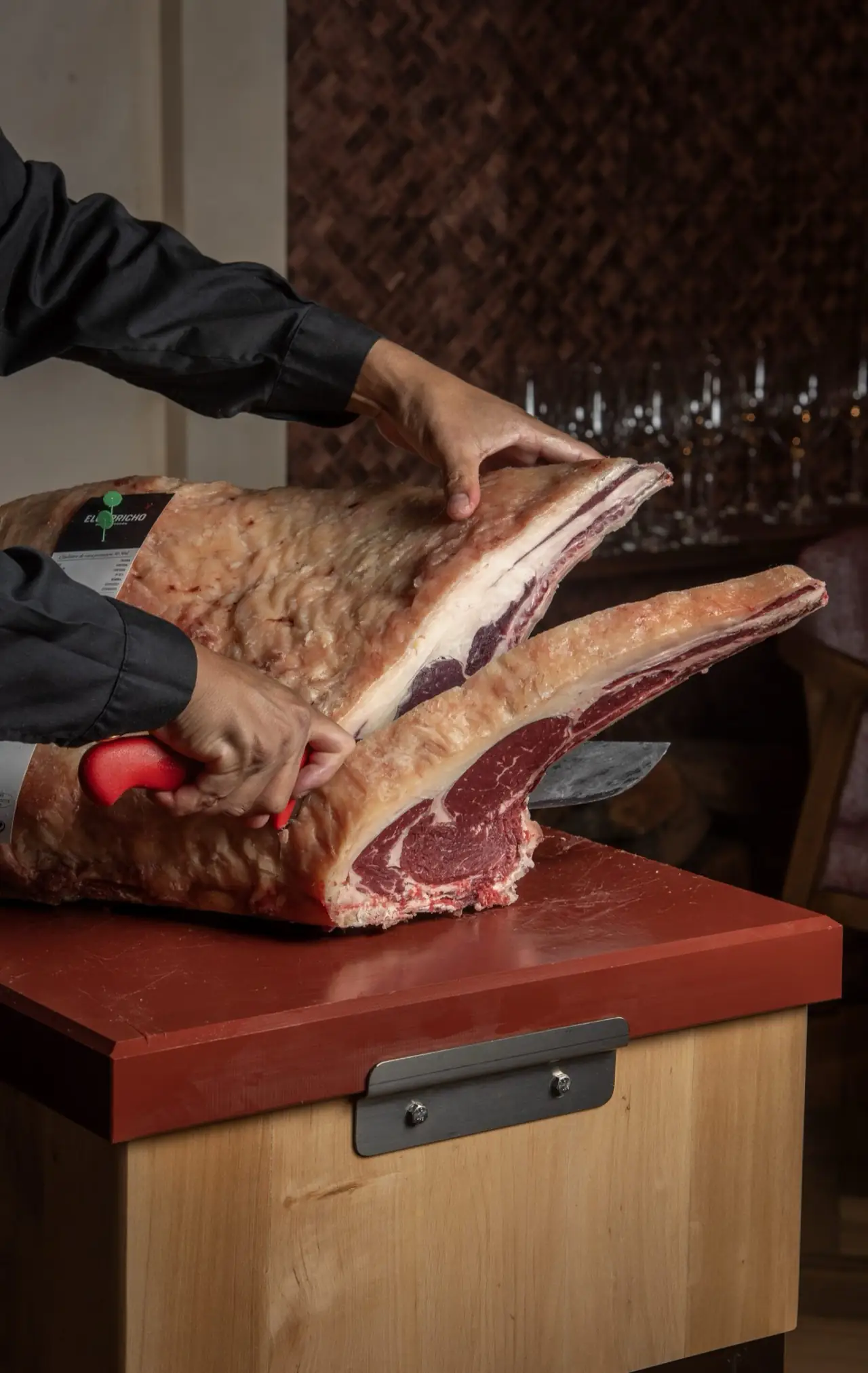 Hands in a black chef jacket trimming a large bone-in dry-aged beef rib with a red-handled knife on a red cutting board