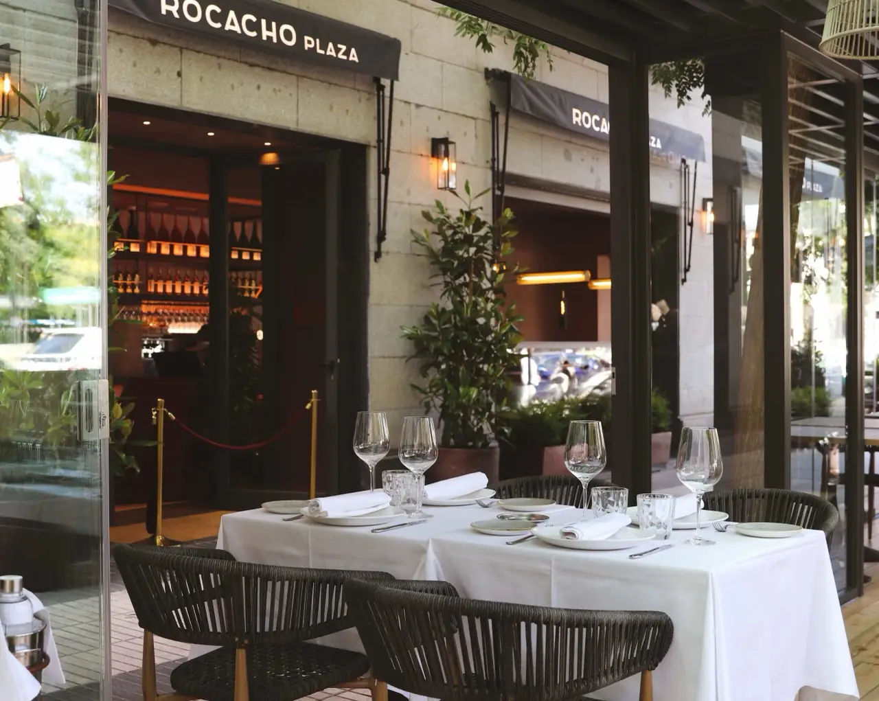 Rocacho Plaza terrace with a white-linen table set with wine glasses and plates, the venue's awning signage and glass-fronted wine wall behind