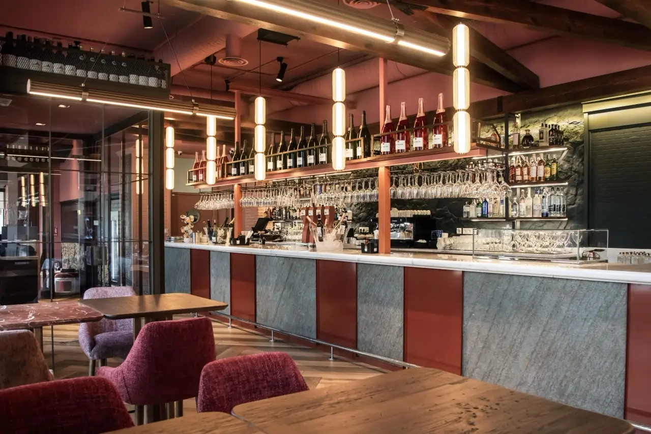 Interior bar with a marble-topped counter, rows of hanging stemware and champagne bottles overhead, terracotta and grey tones with red upholstered chairs
