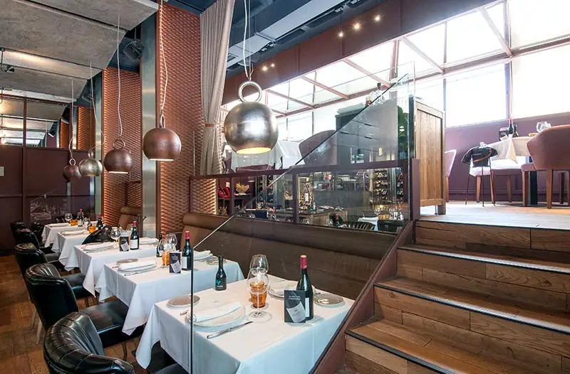 Dining room with white-linen tables set with wine bottles, distinctive silver bell-shaped pendant lamps, and an open kitchen behind a glass partition