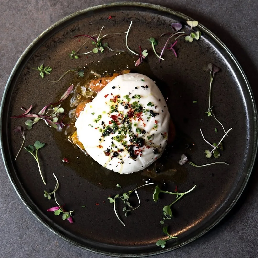 A burrata with herbs and microgreens on a dark plate