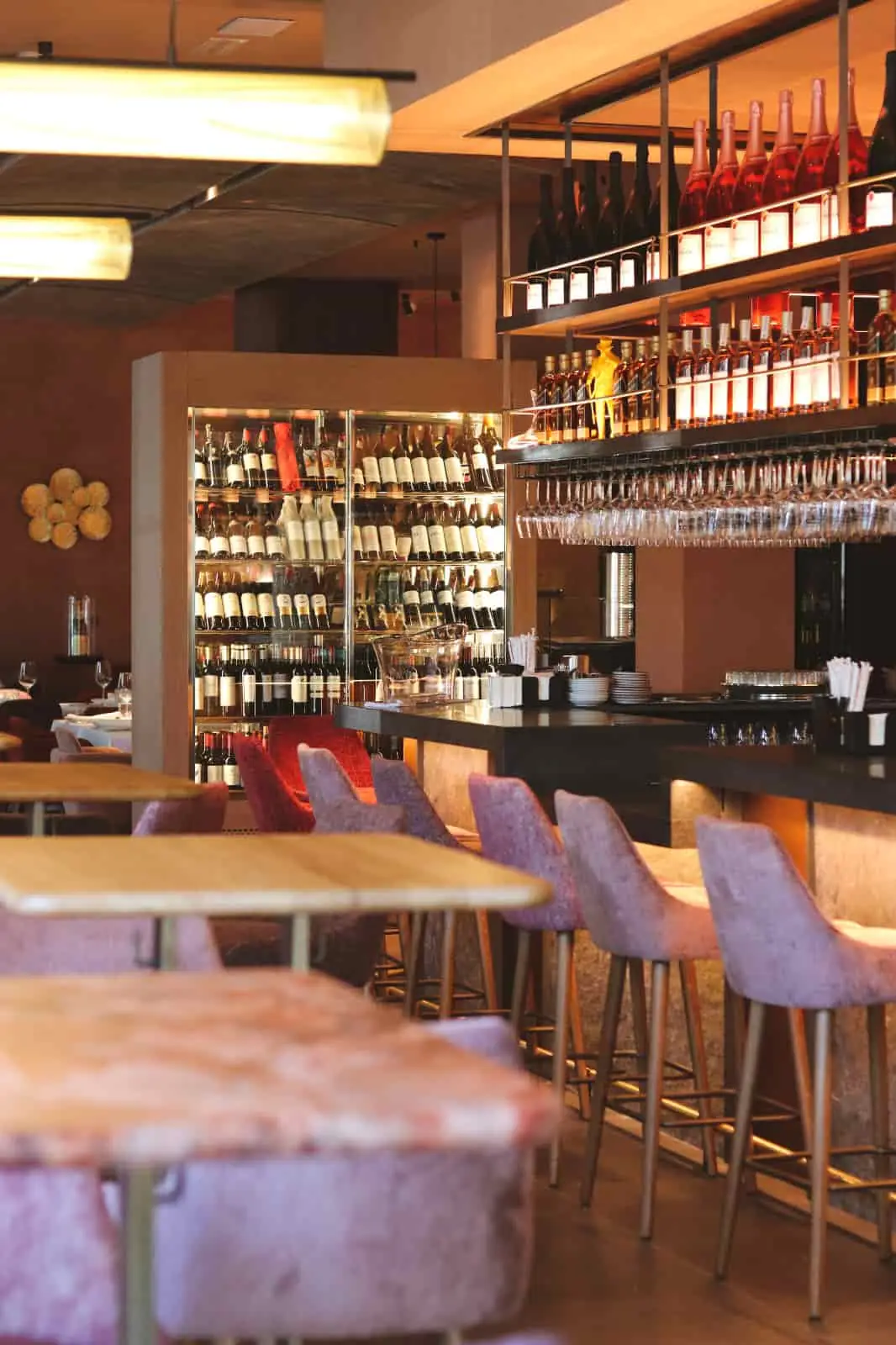 Interior bar with a backlit wine display wall of bottles, hanging wine glasses over a dark bar counter, and violet velvet bar stools beside wooden tables