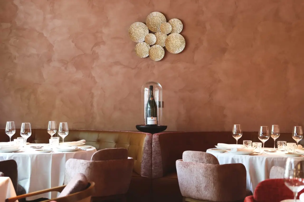 Dining room with terracotta plaster walls, round white-clothed tables set with wine glasses, mauve tub chairs and banquettes, and a champagne bottle under a glass dome on the wall
