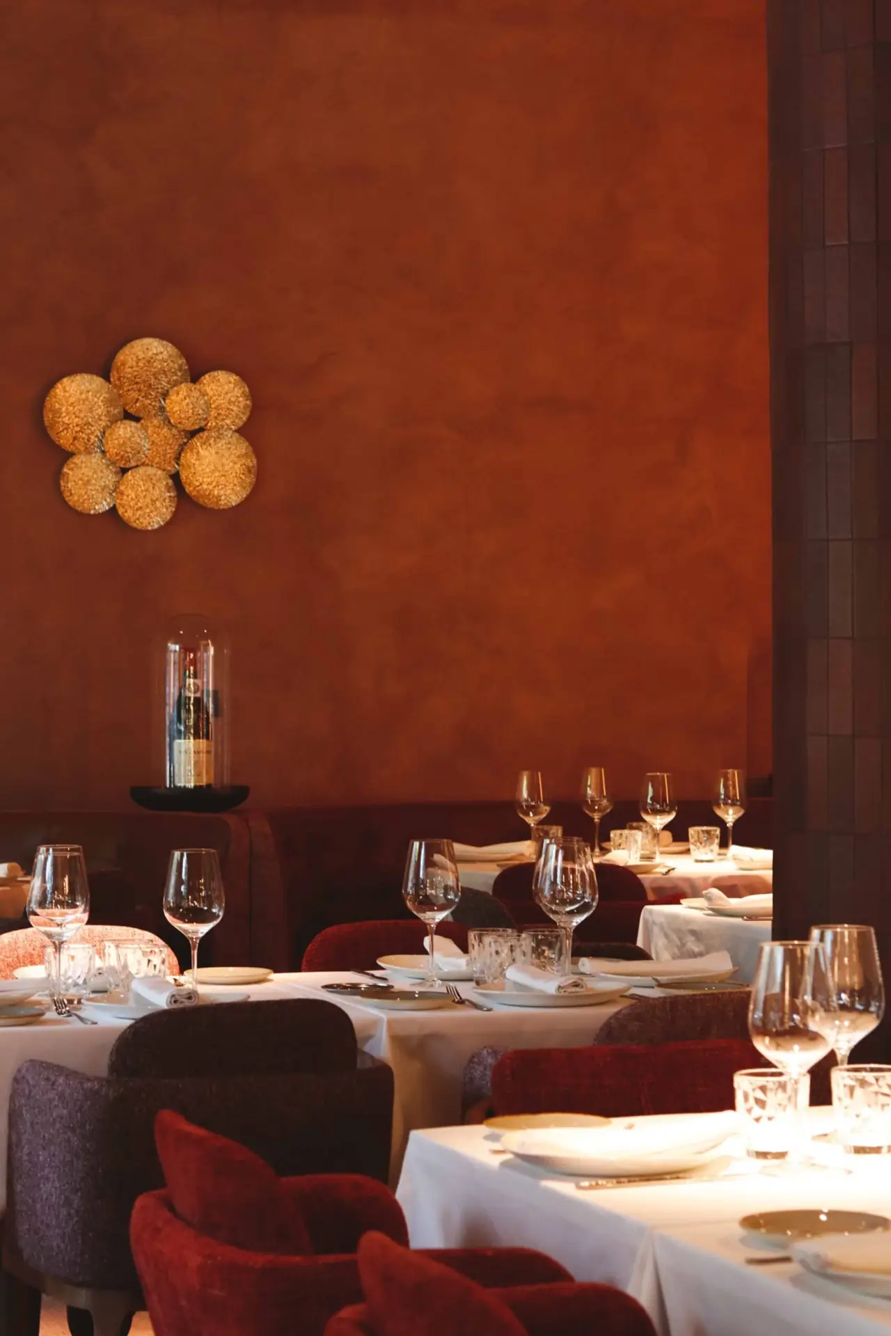 Warm-lit dining room with terracotta walls, rows of round white-clothed tables set with wine glasses and plates, and mauve and red upholstered tub chairs