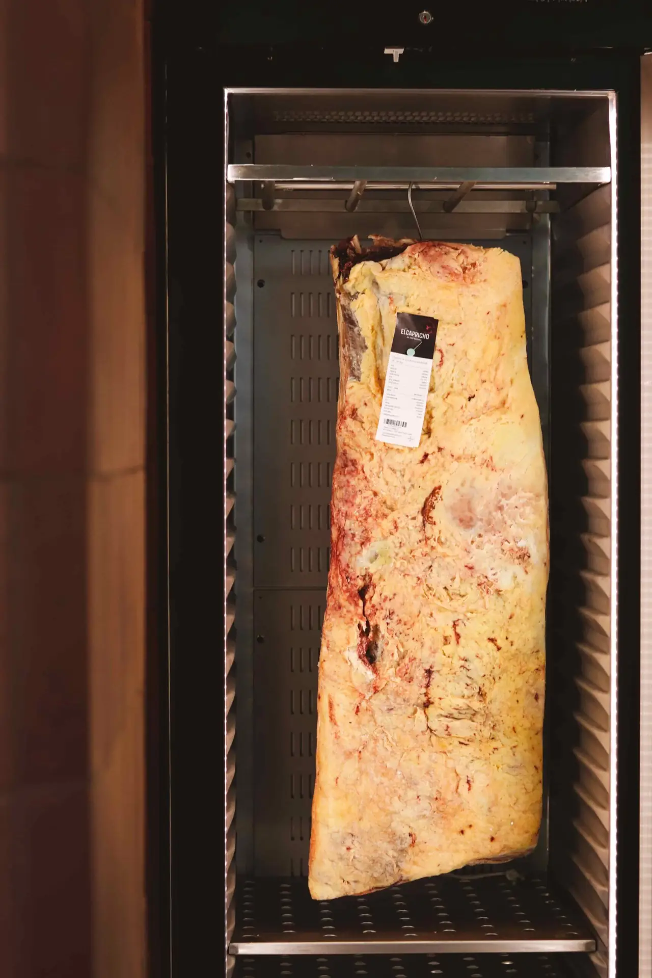A large fat-capped dry-aged beef loin hanging in the venue's stainless-steel aging cabinet, tagged with an El Capricho label