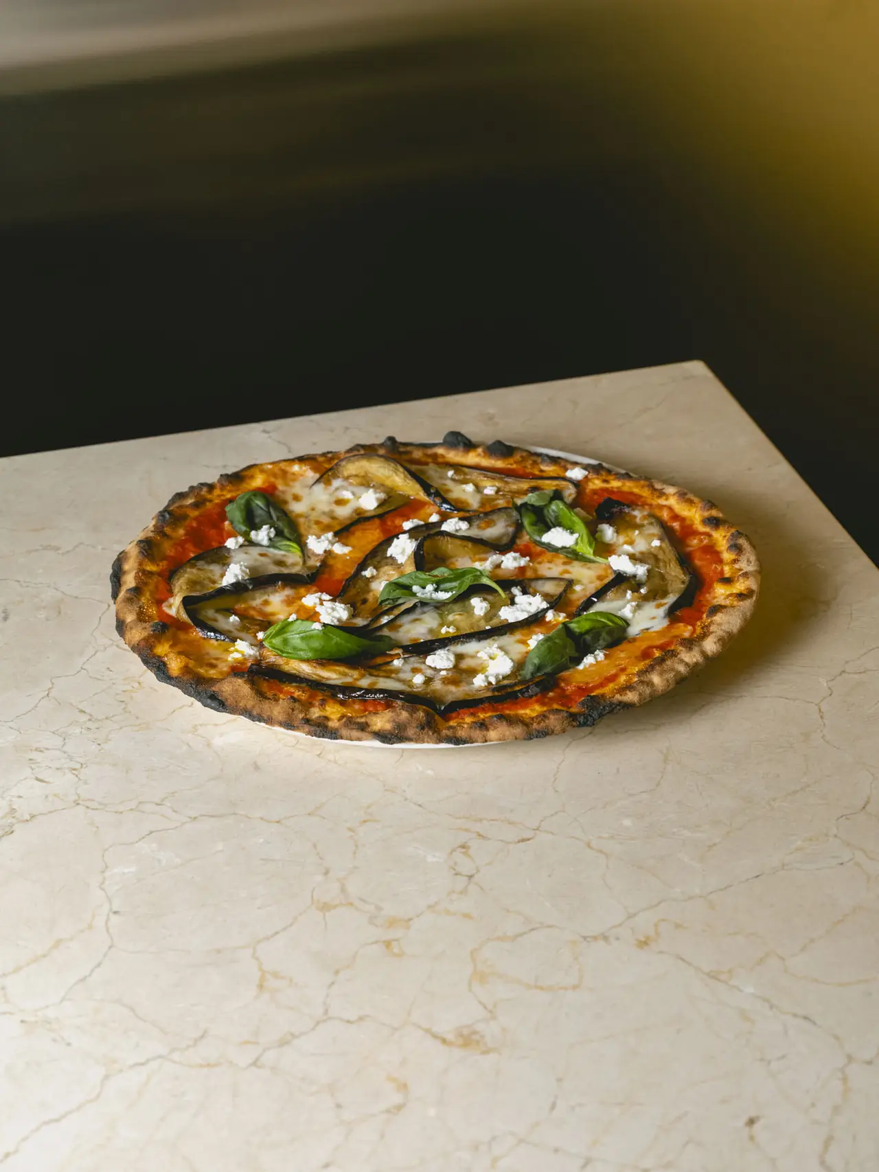 Wood-fired pizza topped with grilled eggplant slices, dollops of soft cheese, and fresh basil leaves on a charred crust, Romo