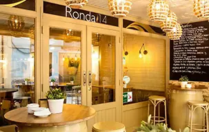 Warmly lit restaurant interior with a rounded wooden counter, white ceramics, brass pendant lamps, glass-partitioned doors under a Ronda 14 sign and a chalkboard menu on the wall