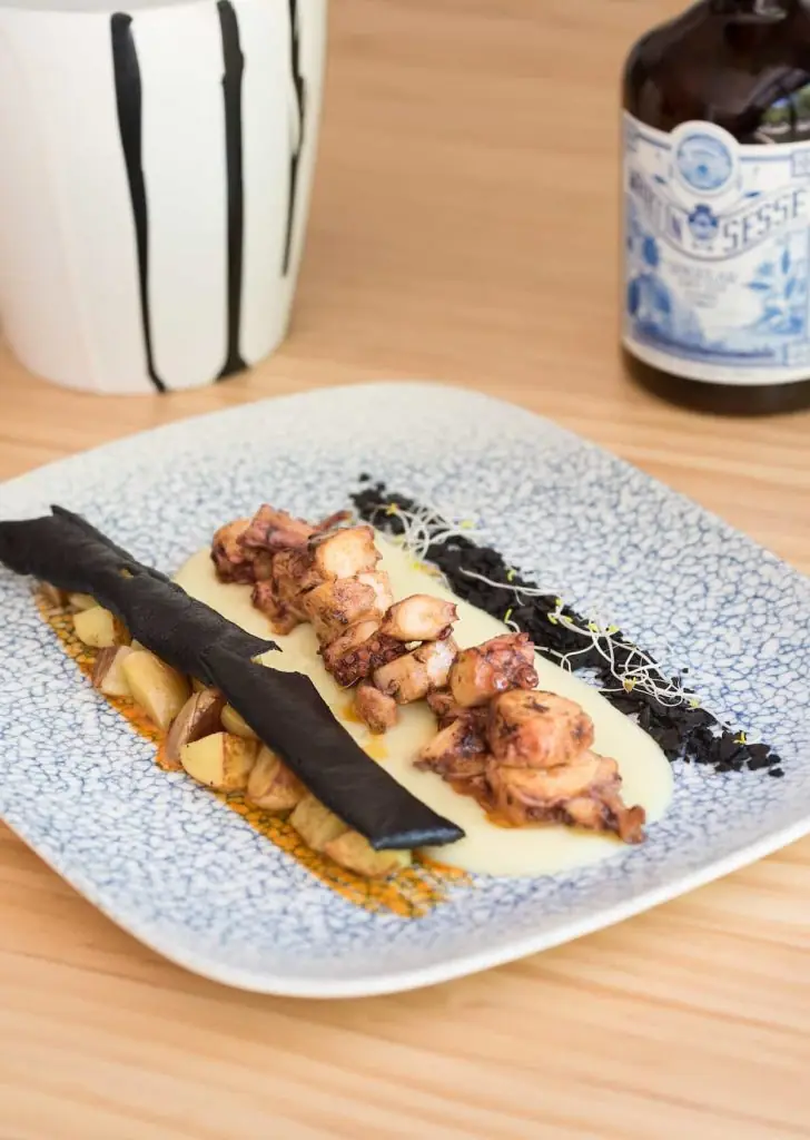 Grilled octopus with roasted potatoes, a crispy squid-ink tuile, and creamy sauce on a blue-patterned plate, served with a bottled beer