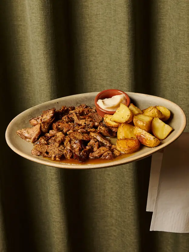 Sliced roast meat with crispy edges served alongside roasted potato wedges and a small dish of aioli, on an oval ceramic plate, at Rooster & Bubbles