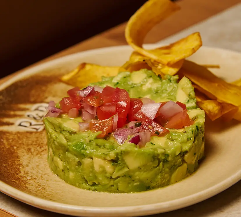 A round mound of chunky guacamole topped with diced tomato and red onion, served with fried plantain-style crisps on a stoneware plate, at Rooster & Bubbles