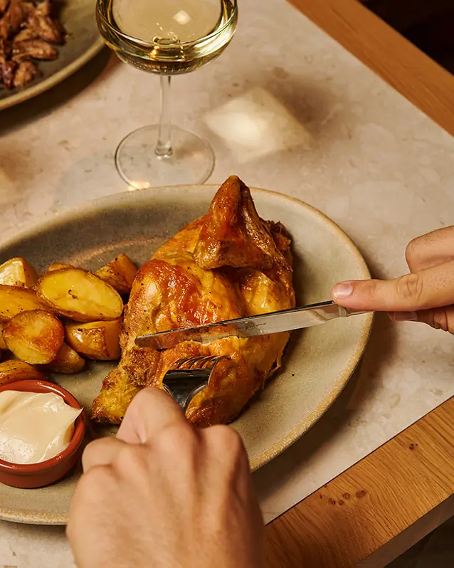 A golden roasted chicken quarter and potato wedges being cut with a knife and fork on a plate, with a small cup of aioli, at Rooster & Bubbles