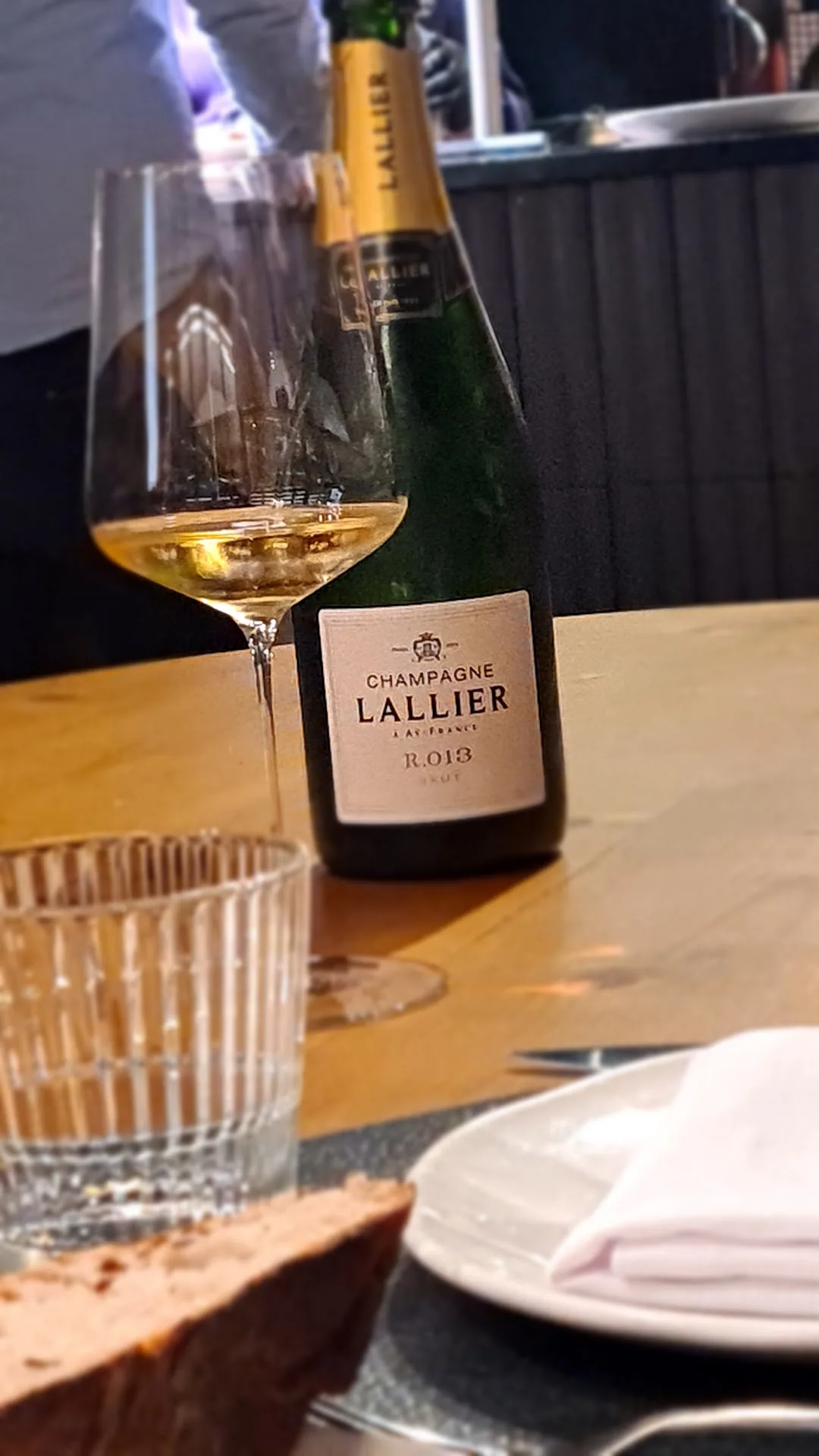 A glass of champagne beside a bottle of Champagne Lallier on a wooden table
