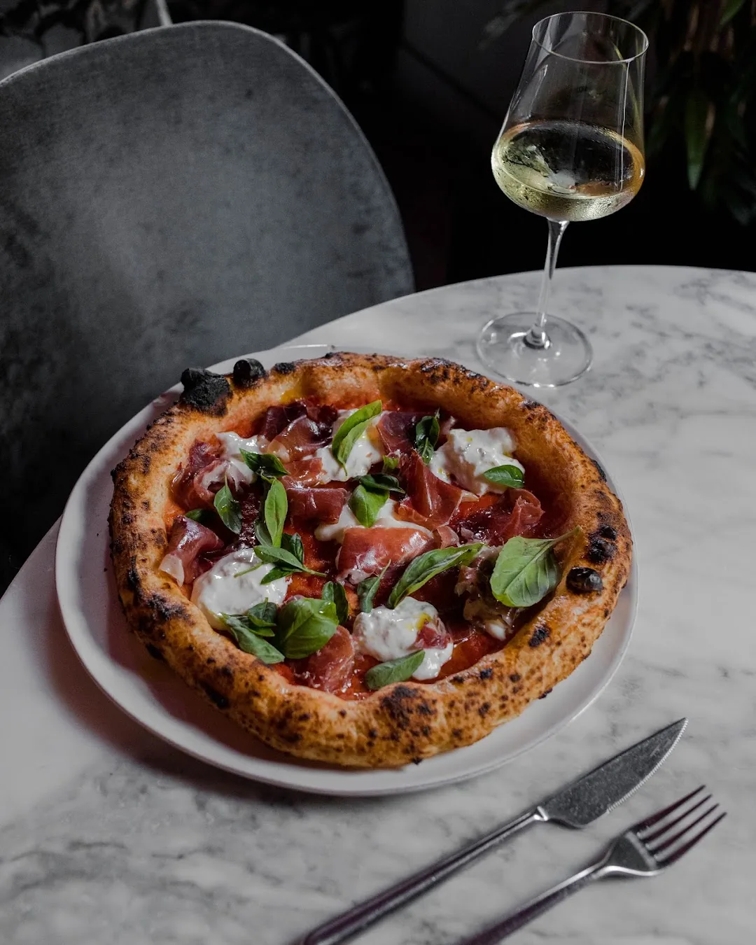 A wood-fired pizza topped with prosciutto, burrata, and basil, plated beside a glass of white wine on a marble table