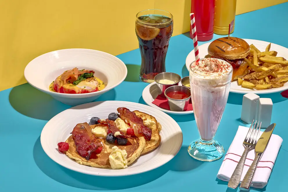 Diner-style spread on a teal table: pancakes with bacon and berries, eggs benedict in a bowl, a strawberry milkshake, a burger with fries and a glass of cola
