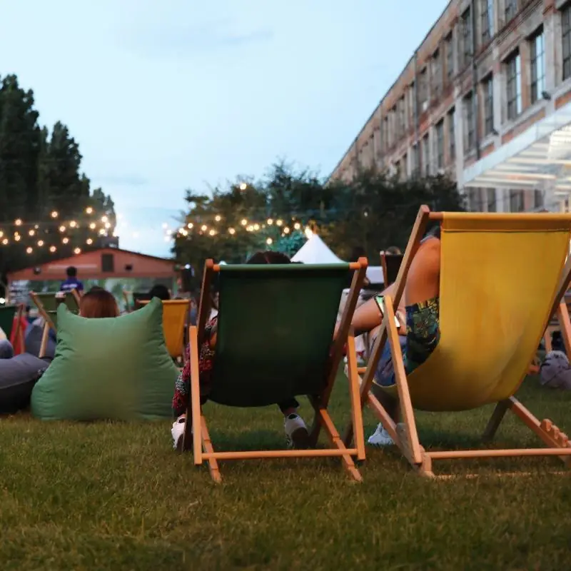 Outdoor garden at dusk with colourful wooden deckchairs and bean bags on a lawn, strung festoon lights and a brick industrial building behind